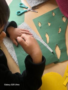 Ein Kind bastelt mit grünem Papier, Filz und Schere auf einem Tisch; warme Farben, kreative Atmosphäre.