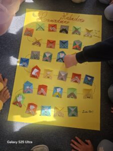 Gelbes Plakat mit handgemachten Umschlägen in Reihen als Ramadan-Kalender, umgeben von Kinderhänden auf grauem Teppich. Helle, fröhliche Atmosphäre.