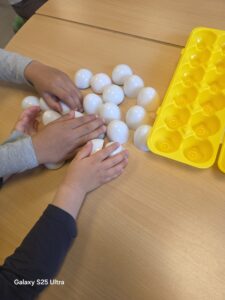 Kinderhände greifen nach weißen Plastikeiern auf einem Holztisch, neben einer gelben Eierschachtel.
