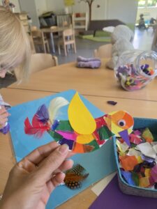 Bastelarbeit eines bunten Papiervogels mit Federn und Wackelauge, gehalten von einer Hand in einem Kita-Raum mit Bastelmaterial auf dem Tisch.