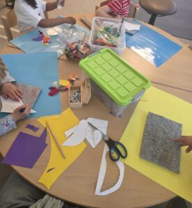 Kinder basteln an einem Tisch mit buntem Papier, Scheren und Kleber. Figuren und Materialien sind lebendig und farbenfroh verteilt.
