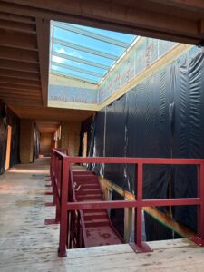 Blick in halbfertigen Bau mit Holzdecke, Oberlicht und rotem Treppengeländer. Schwarze Abdeckplanen verhüllen seitliche Wände.