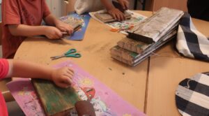 Kinder schleifen Holzstücke auf einer Tischoberfläche, umgeben von Scheren und bedruckten Unterlagen. Die Stimmung ist kreativ und arbeitsintensiv.