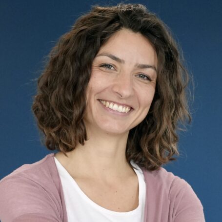 Frau mit lockigem Haar lächelt, trägt weißes Shirt und rosa Cardigan. Blauer Hintergrund, freundliche und entspannte Stimmung.