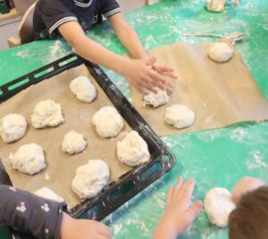 Kinder rollen Teigkugeln auf einem bemehlten Tisch; Backblech und Backpapier mit weiteren Teigkugeln. Verspielte und kreative Stimmung.