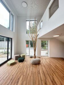 Moderner Innenraum mit hohen weißen Wänden, großen Fenstern, Holzboden. Sitzkissen in Pastellfarben und ein dekorativer Ast in der Mitte. Rustikales, helles Design.
