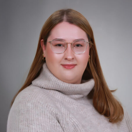 Porträt einer Person mit langen Haaren, Brille und hellem Rollkragenpullover vor grauem Hintergrund. Die Stimmung ist neutral.