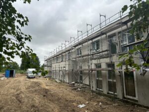 Zwei­stöckiges Gebäude mit Gerüst, im Bau­zustand. Erdiger Boden im Vordergrund, Bäume und bewölkter Himmel im Hintergrund.