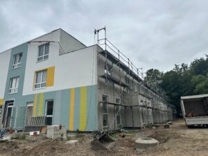 Mehrstöckiges Gebäude im Bau mit Gerüst, teils bunte Fassade. Baustellenmaterialien und LKW im Vordergrund, bewölkter Himmel.