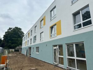 Mehrstöckiges Gebäude mit weißen, gelben und mintgrünen Fassadenelementen. Unbefestigter Boden und Baum im Hintergrund.