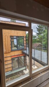 Blick durch ein großes Fenster auf ein modernes, mit Holz verkleidetes Gebäude und eine Metallveranda, umgeben von Bäumen bei bewölktem Himmel.