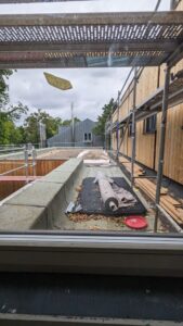 Blick durch ein Fenster auf eine Baustelle mit Gerüst, Baumaterialien und teils verlegtem Kies auf einem Flachdach; bewölkter Himmel.