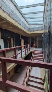 Ein halbwegs fertiggestelltes Treppenhaus mit roten Geländern, schwarzer Plastikfolie an den Wänden und einem Glasdach für Tageslicht.