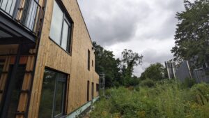 Holzverkleidetes Gebäude mit großen Fenstern links, daneben dichter Bewuchs und Bäume unter bewölktem Himmel. Rechts sind metallische Strukturen zu sehen.