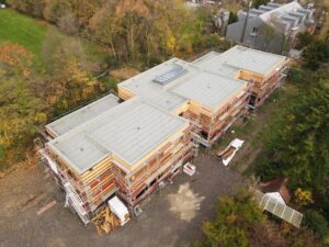 Luftaufnahme eines im Bau befindlichen Gebäudes mit Flachdach und Gerüsten. Umgeben von Bäumen in Herbstfarben, ruhig und strukturiert.