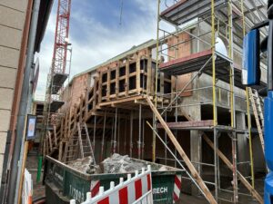 Rohbau eines Gebäudes mit Gerüsten und Baukran, Ziegelwände und Betonflächen, daneben ein Schuttcontainer und Absperrgitter. Helle, wolkige Stimmung.