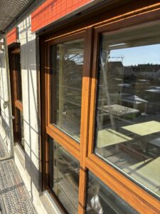 Braunes Holzfenster in einer Hauswand auf einer Baustelle, mit Baugerüst im Vordergrund und sonnigem Wetter.