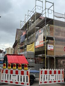 Ein mit Baugerüst umgebenes Gebäude, davor Absperrungen mit blinkenden Lichtern und Baumaterialien; bewölkter Himmel im Hintergrund.