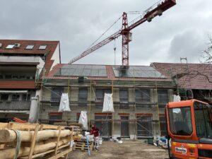 Gebäudesanierung mit Baugerüst, Solarpanels auf dem Dach, Kran im Hintergrund, Baumaterialien und ein kleiner Bagger im Vordergrund; bewölkter Himmel.
