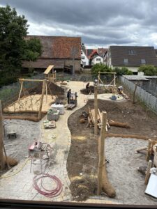 Baustelle für einen Spielplatz mit Holzkonstruktionen, Kieswegen und Erdhügeln. Bäume und Gebäude im Hintergrund, bewölkter Himmel.