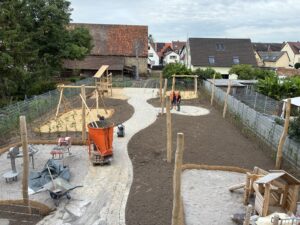 Spielplatz im Bau mit Holzspielgeräten, sandigem Boden, zwei Personen in Baukleidung, umliegende Häuser und Bäume; helle, bewölkte Atmosphäre.