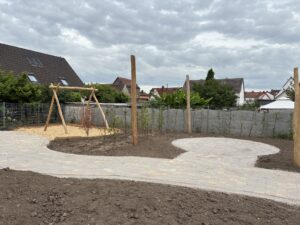 Spielplatz mit Holzschaukel auf Mulch, umgeben von gepflastertem Weg und kahlem Boden; im Hintergrund Wohnhäuser und bewölkter Himmel.