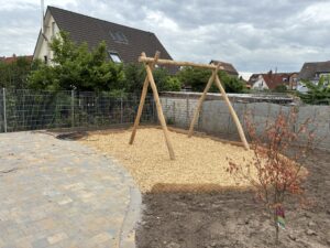 Holzschaukel auf Kiesfläche in kleinem Garten, umgeben von Metallzaun, gepflasterter Terrasse und einigen Büschen; bewölkter Himmel.