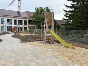 Spielplatz im Bau mit Rutsche, Klettergerüst und gepflastertem Weg. Hintergrund: Wohnhaus mit Solarzellen, Baukran, grüne Bäume.