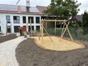 Gartenbaustelle mit Holzspielgerüst, Holzschnitzelboden, Gebäuden im Hintergrund und einem Arbeiter auf gepflastertem Weg bei bedecktem Himmel.