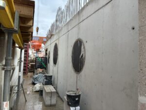 Baustelle mit Betonwand, zwei runden Aussparungen, Gerüsten und Bauzubehör. Graue und orangefarbene Elemente dominieren die Szene.