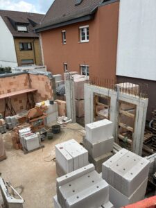 Baustelle zwischen Wohnhäusern; unverputzte Mauern, Ziegelblöcke, Mischmaschine. Teilweise fertige Betonwände, klare Tageslichtstimmung.