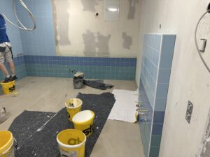 Baustelle in einem Raum mit hellblauen Fliesen an den Wänden, Eimern und Werkzeugen auf dem Boden, eine Person arbeitet auf einem gelben Eimer.
