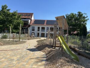 Spielplatz mit Holzklettergerüst und grüner Rutsche vor einem Gebäude mit Solarpanelen auf dem Dach. Gepflasterter Weg und Bäume im Hintergrund.