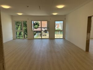 Leerer Raum mit hellem Laminatboden, große Fenster mit Blick ins Freie, an der Decke runde Leuchten. Helle, neutrale Atmosphäre.