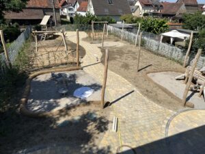Ein neu angelegter Spielplatz mit Holzgerüsten, Sandflächen und gepflasterten Wegen. Ruhige, aufgeräumte Atmosphäre bei sonnigem Wetter.