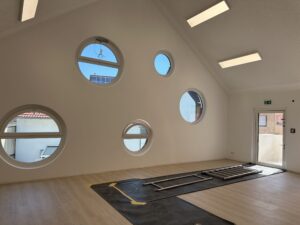 Heller Raum mit schräger Decke, runden Fenstern, Holzfußboden und Leiter. Fenster bieten Ausblick auf Dächer. Moderne und minimalistische Gestaltung.