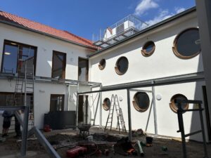 Innenhof eines Gebäudes im Bau, weiße Fassade mit runden Fenstern, Leitern und Baugeräte, blauer Himmel; klare, helle Atmosphäre.