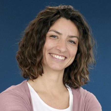 Eine lächelnde Person mit lockigem Haar vor blauem Hintergrund, trägt ein weißes Oberteil und eine rosa Strickjacke.